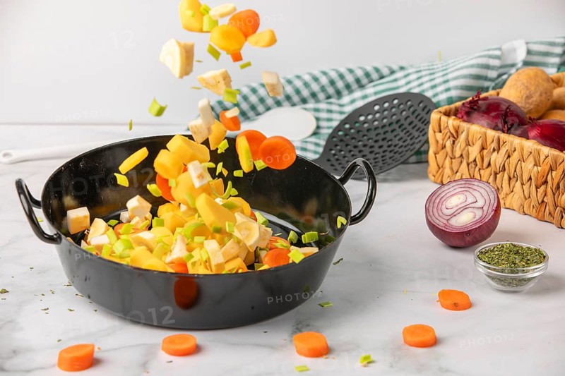 Fresh soup vegetables fall into a cooking pot next to ingredients and kitchen utensils