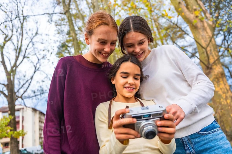 Three smiling sisters or friends discovering the functions of an old camera while enjoying a lovely family leisure time together in an urban park during autumn
