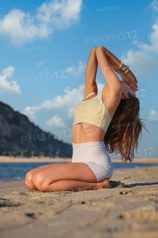 Woman performs Vajrasana Thunderbolt Pose with backbend on a sandy beach during sunset, kneeling with arms raised overhead