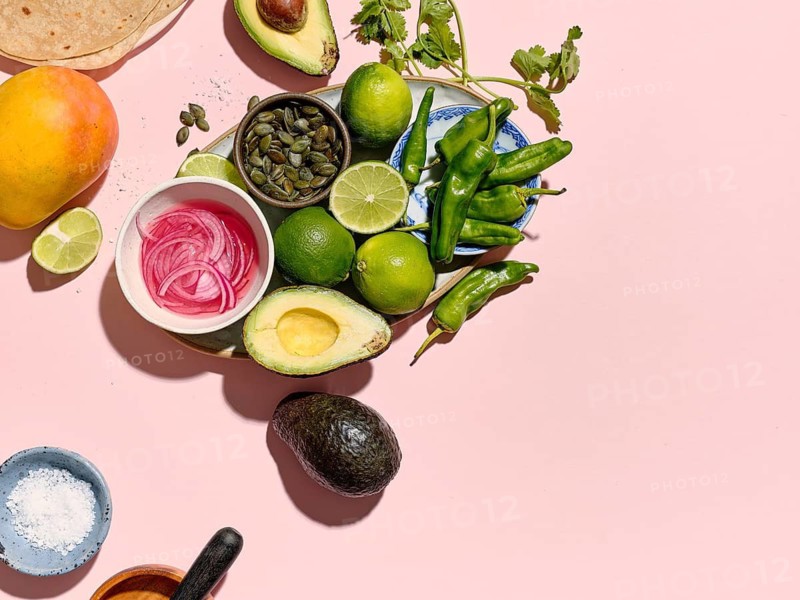Top view of a vibrant taco preparation scene on a pink background. Ingredients like avocados, limes, a mango, green chilies, pumpkin seeds, red onions, salt, and coriander are scattered artistically. Ideal for culinary guides and recipe layouts
