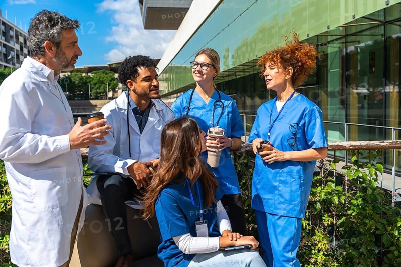 Diverse doctors and nurses chat and sip coffee on a sunny break outside a modern hospital, smiling and collaborating informally to discuss patient care and team plans