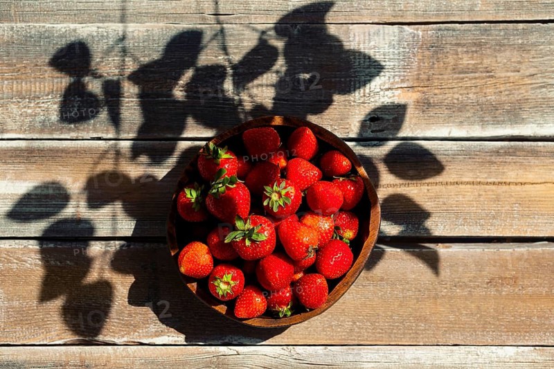 Top view of bowl with ripe strawberries on wooden table. Shadows from nearby leaves create patterns on tabletop. Bright afternoon light highlights freshness of berries in bowl