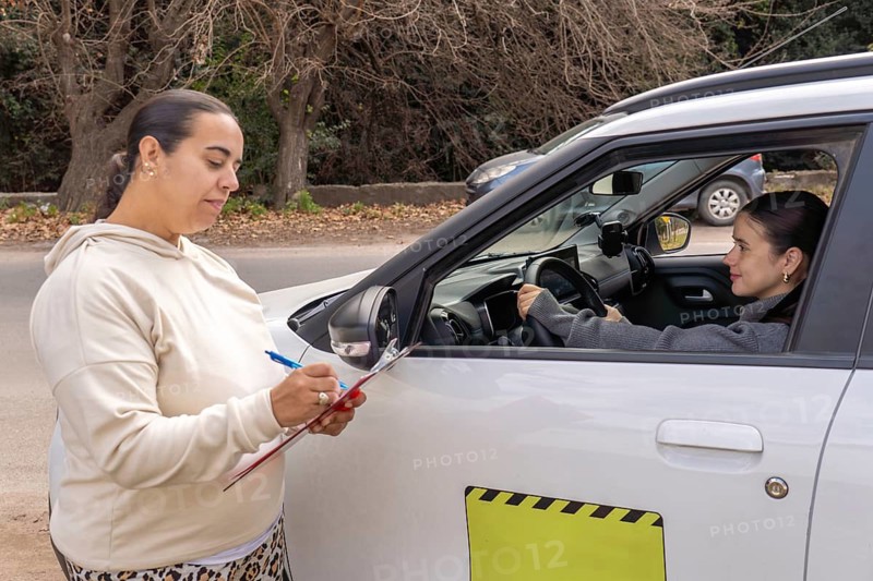 Female driving test examiner writing on clipboard outside car while smiling candidate sits behind the wheel, exam evaluation concept