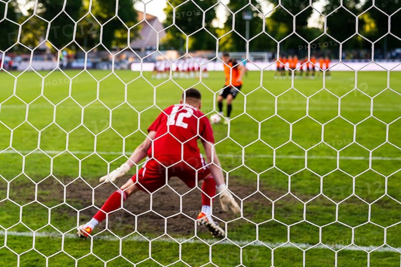 View through the net of a soccer goalkeeper in a red kit preparing to defend a penalty kick during a football match in Glogow, Poland