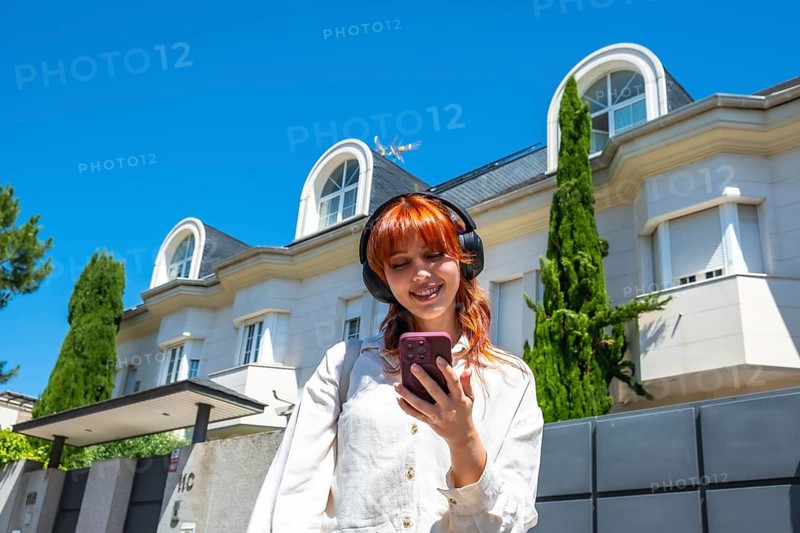 Young woman with red hair wearing wireless headphones and smiling, looking at her smartphone while walking outdoors on a sunny day in a residential area