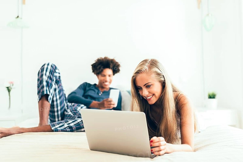 A young woman is using a laptop while a man relaxes with a smartphone on a bed. They are in a light-filled, comfortable bedroom setting