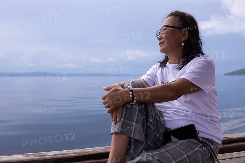 A mature woman sits contentedly on a wooden deck on Kri Island, looking out over the calm waters of the Raja Ampat Archipelago in West Papua, Indonesia. She appears relaxed and introspective, enjoying the serene natural environment