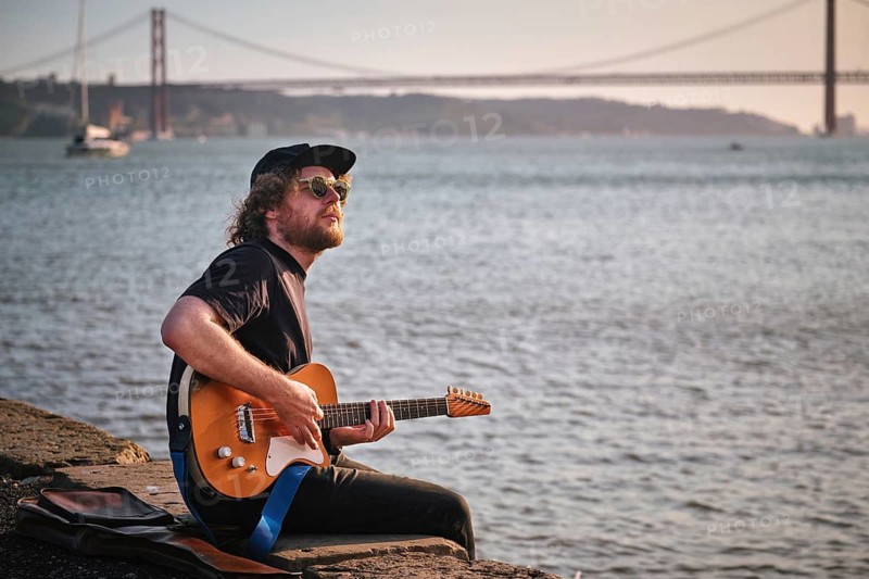 Hipster street musician in black playing electric guitar in the street sitting on pier embankment on sunset