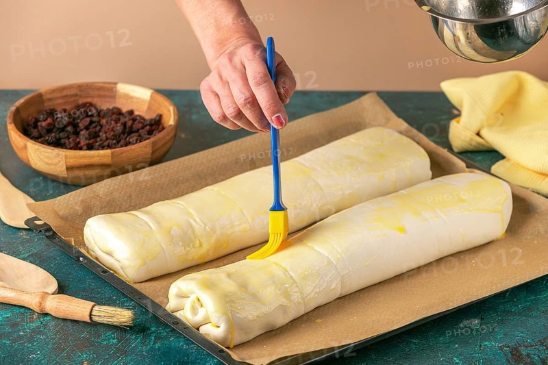 Hand spread strudel with egg on baking tray next to bowl with raisins