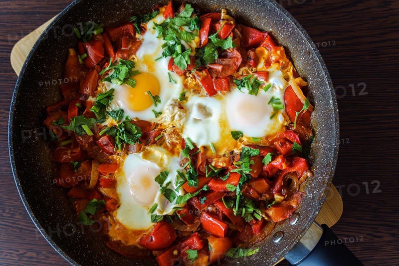 Ready-to-eat shakshouka with eggs poached in sauce of tomatoes, olive oil, peppers, onion and garlic. Traditional mediterranean cuisine. Concept of keto meal, FODMAP recipe and low carb diet. Closeup, table top view, horizontal. A skillet filled with shakshuka, a dish of eggs cooked in a mixture of tomatoes, onions and red peppers, garnished with fresh herbs. The vibrant colors and rustic presentation highlight a freshly prepared Mediterranean meal