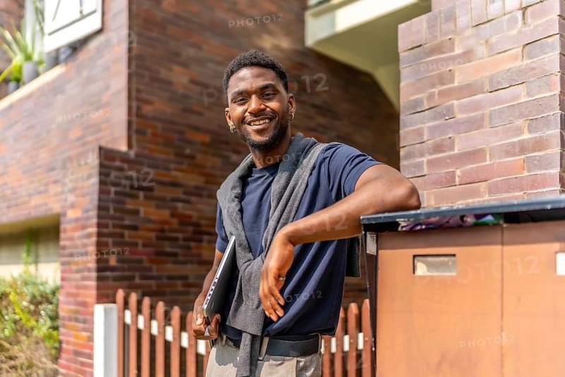 African young businessman leaning in a residential house entrance carrying a laptop and smiling at camera