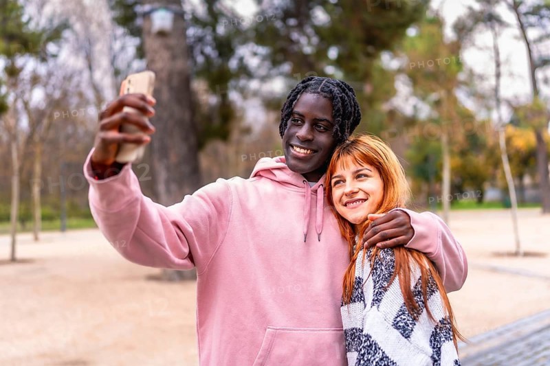 Young multiracial couple smiling and capturing a joyful selfie with a smartphone while enjoying a sunny day in the park