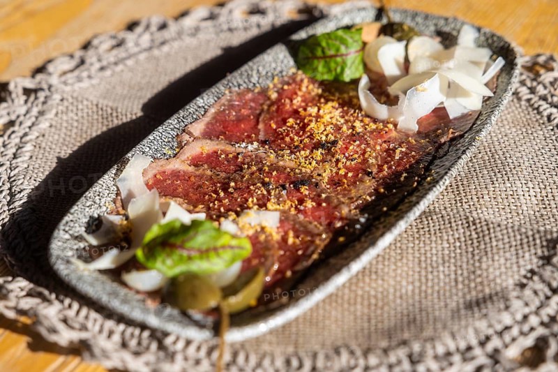 Freshly prepared venison carpaccio adorned with grated parmesan, spices, and a touch of fresh herbs, presented on a textured, dark plate, adding a rustic charm under natural daylight