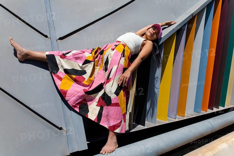 A woman in a vibrant, colorful outfit contrasts strikingly with the sleek, geometric lines of modern architecture. Her pose adds dynamism to the urban scene