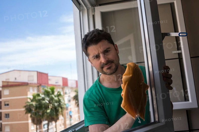 A man engages in household chores by cleaning a window in a modern urban apartment. The scene captures daily life in the city, with a focus on home maintenance tasks