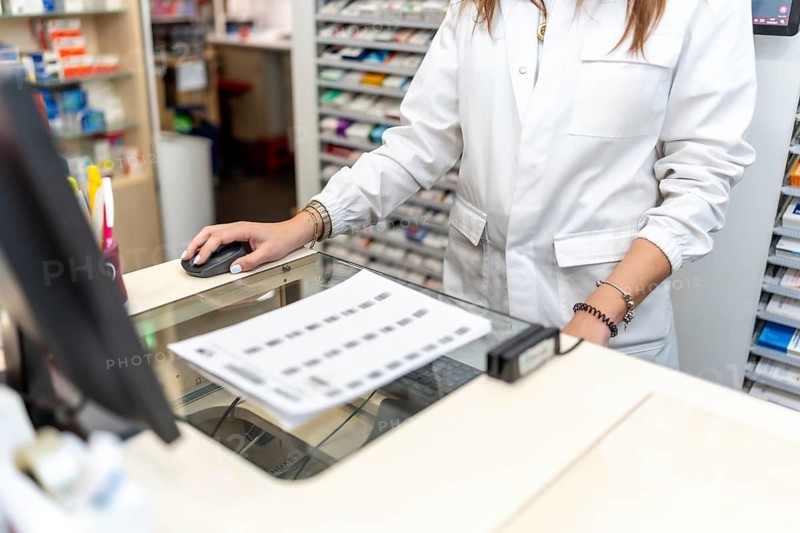 Pharmacist in a lab coat skillfully using a computer mouse on the counter, managing prescriptions and medication orders in a busy retail drugstore environment
