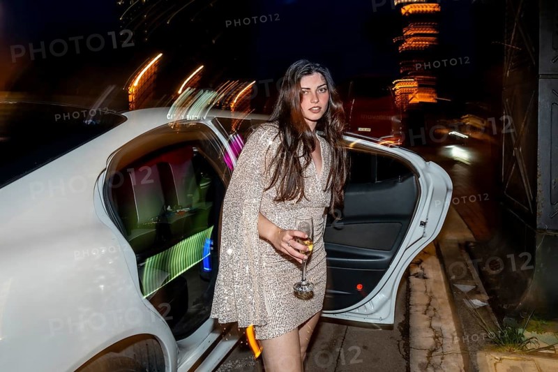 Glamorous woman with long dark hair, wearing a sparkling gold dress, stepping out of a luxurious white limousine at night while holding a glass of champagne, celebrating an elegant event