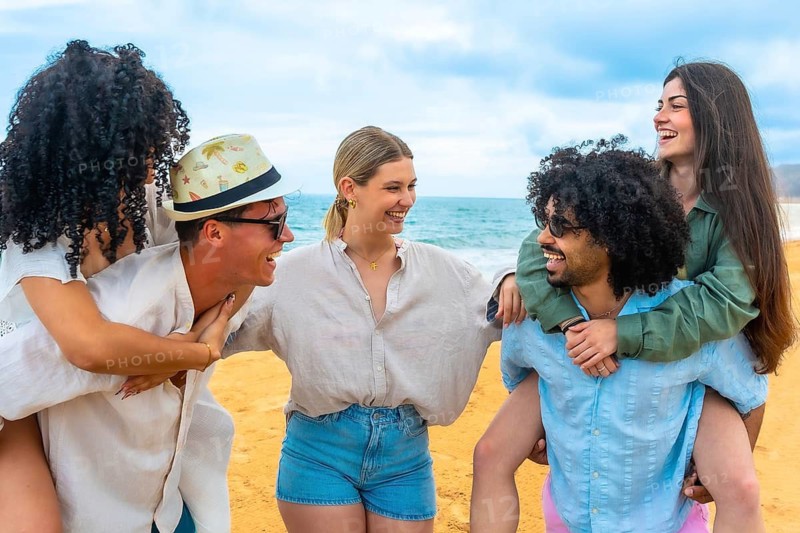 Diverse group of smiling young friends engaging in piggyback rides and having fun on a sunny beach, celebrating friendship, youth, and summer vacation by the sea