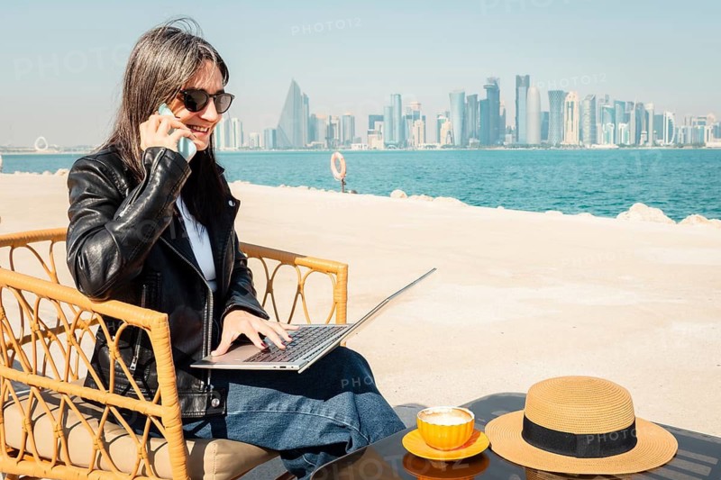 Happy business woman drinking coffee using laptop and smartphone chat. West Bay skyline background digital nomad working remotely in outdoor cafe. Middle East city urban professional