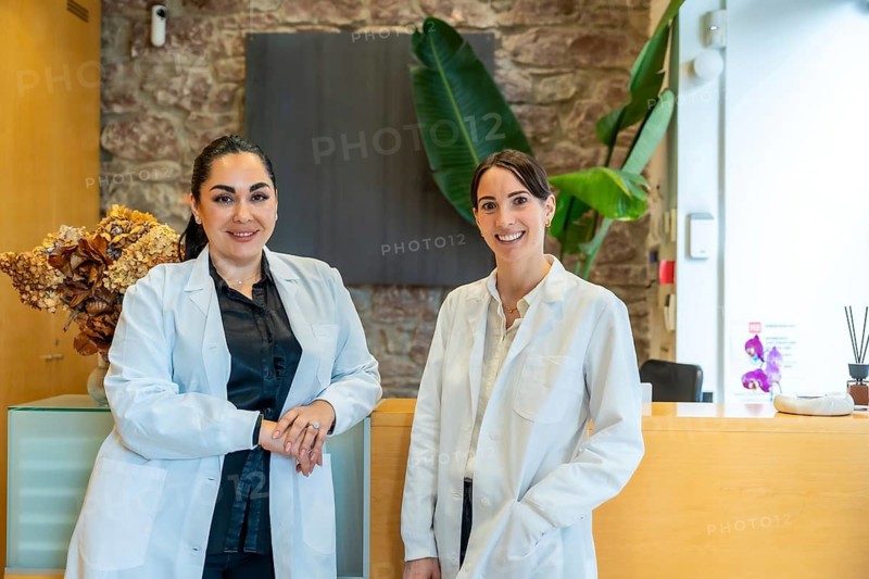 Two smiling female medical professionals wearing lab coats standing in a contemporary clinic or spa reception area, representing healthcare, wellness, and medical services