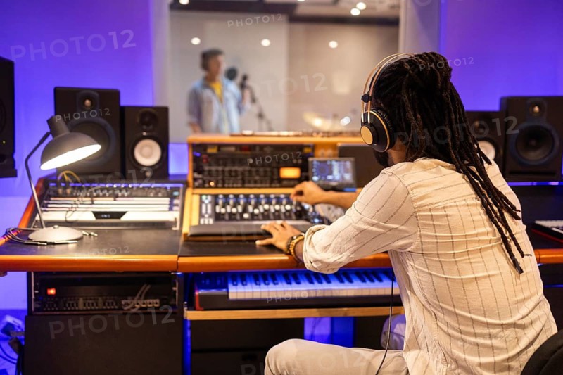 An audio engineer wearing headphones operates a mixing console in a modern recording studio, capturing a vocalist performing in the soundproof booth