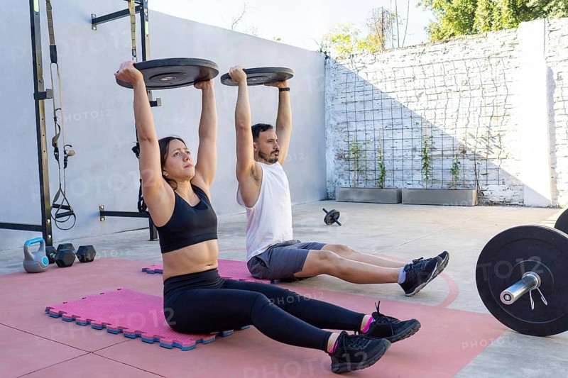 Young hispanic man and woman lifting weights while sitting on mats in a backyard gym, embracing a healthy lifestyle together and enjoying their workout in the summer sunlight