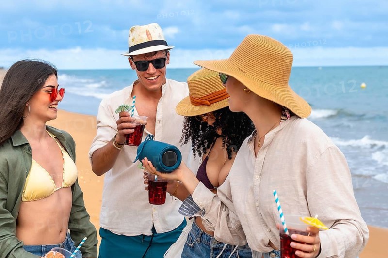 Young friends chilling on a sunny beach with colorful cocktails and a portable speaker, enjoying music, laughter, and carefree summer vibes by the sea and sand