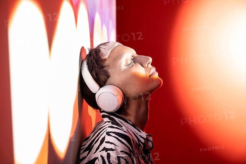 A young trendy woman, eyes closed, leans against a wall wearing stylish headphones. The warm background light enhances the modern and relaxed atmosphere of the scene