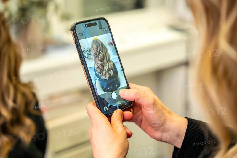 Hands holding smartphone capturing a young woman's long wavy hair in a salon, highlighting successful hairstyling and beauty transformation for social media promotion