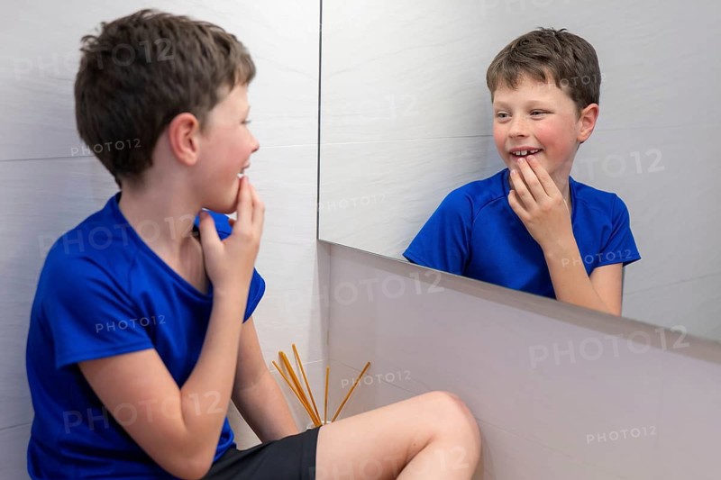 A young boy in a blue shirt smiles at his reflection, touching the gap in his teeth where a milk tooth recently fell out