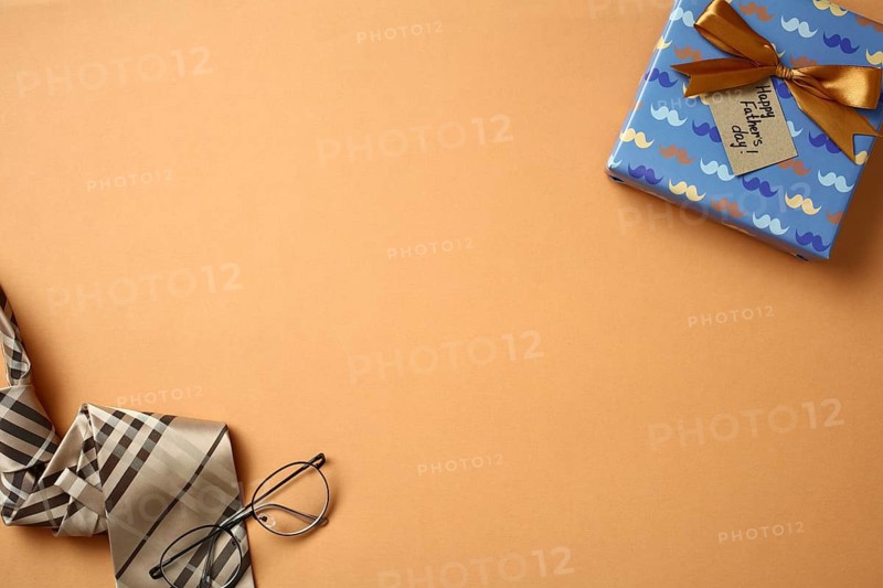 Top view of a stylish Father's Day setup with a wrapped gift featuring a blue and gold ribbon, a Happy Father's Day tag, elegant glasses, and a striped tie, all neatly arranged on a orange background