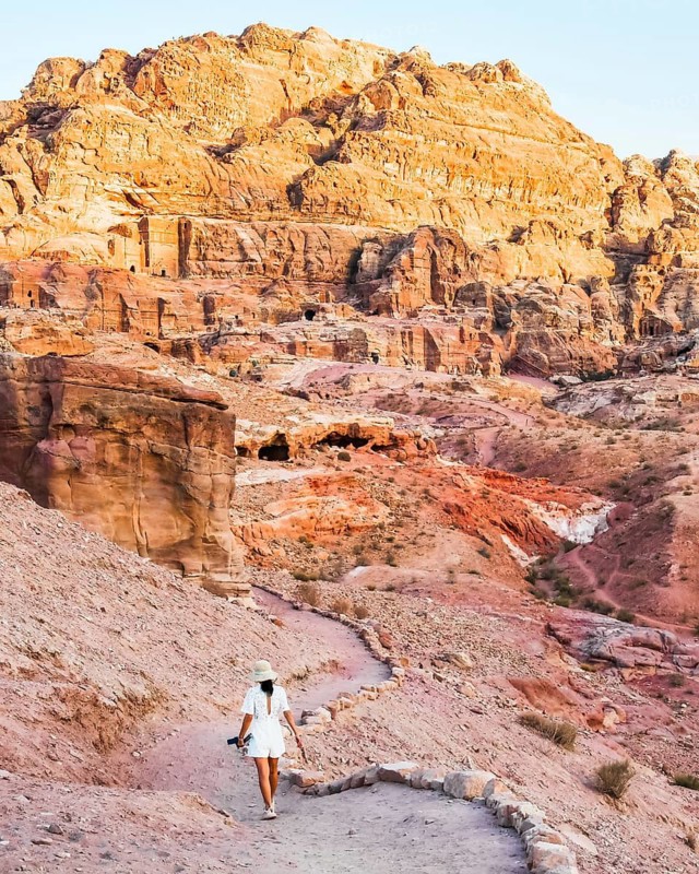 Solo traveler walking along a winding path through Petra's sandstone landscape and ancient Nabataean rock-cut structures at golden hour. Iconic Jordan desert heritage site and Middle East travel destination