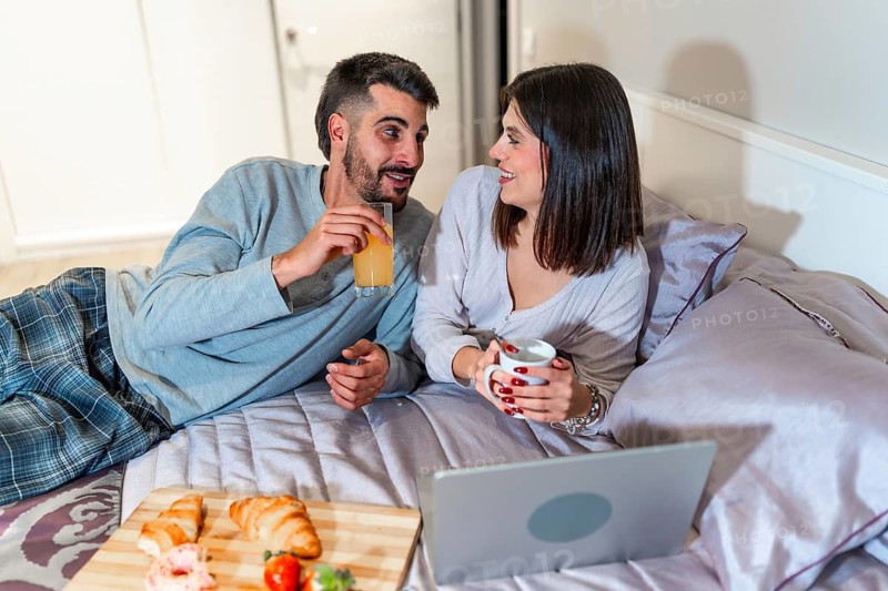 Young couple lying in bed, having breakfast and watching something on their laptop, spending quality time together