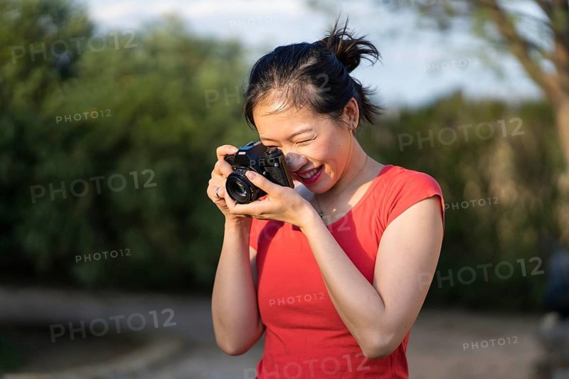 Asian woman in a red shirt capturing moments outdoors with a digital camera. Her joyful expression and natural surroundings create a vibrant and inspiring scene for photography lovers