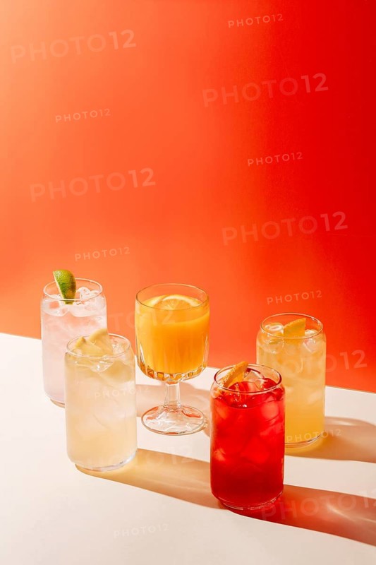 Five assorted lemonades displayed on a surface, set against a bright orange backdrop. Each glass features different citrus accents, offering a refreshing visual appeal