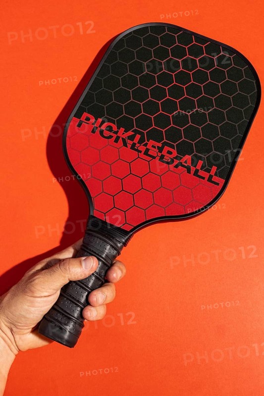 Cropped unrecognizable hand grips a pickleball paddle against a vivid orange background, highlighting the energetic and modern look of the sports equipment in a studio setting