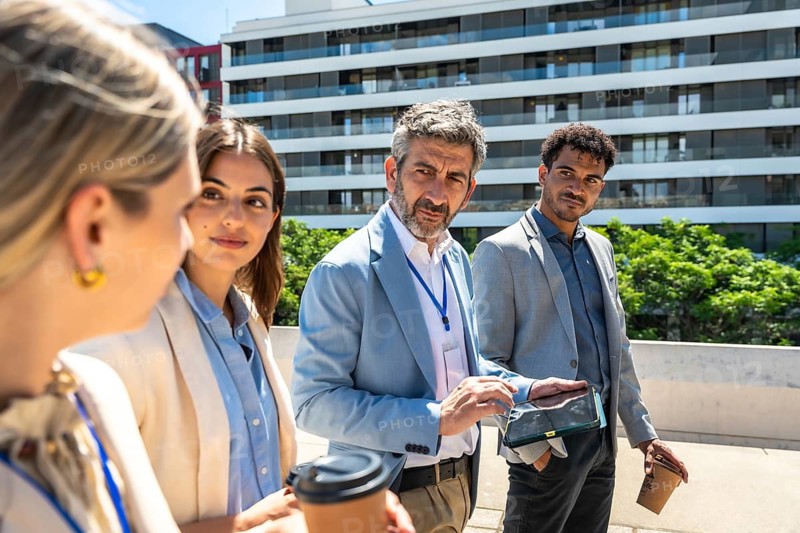 Diverse business professionals standing outdoors, discussing work and reviewing information on a digital tablet while holding coffee, collaborating on a project in an urban setting