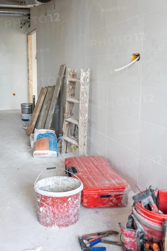 Empty room showing renovation progress with a dirty red bucket, toolbox, ladder, construction materials, and pipes, indicating an ongoing building or installation project