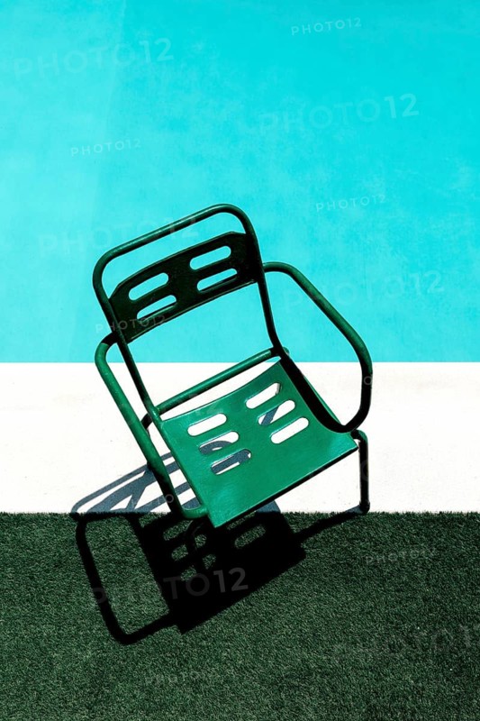 Top view of a green metal chair casting a shadow on artificial grass beside a pool. The vibrant blue water and sunny ambiance evoke a perfect summer day