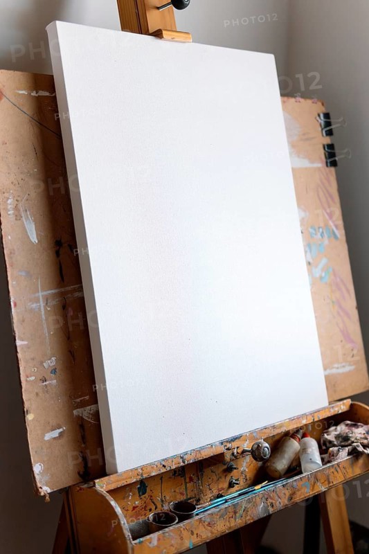A clean, blank canvas rests on a well-used wooden easel covered in paint splatters. Art supplies sit nearby, waiting for the next creative masterpiece to take shape