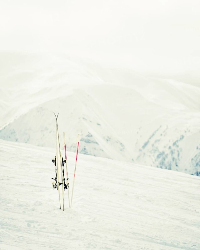 Pair of white skis with poles planted in snow on mountain slope with alpine peaks in background, concept of winter sport, ski holiday, rentals, quality gear and day off from slopes