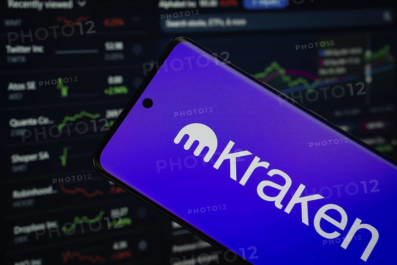 kraken logo is displayed on a smartphone screen, with a stock graph webpage visible on a computer screen in the background