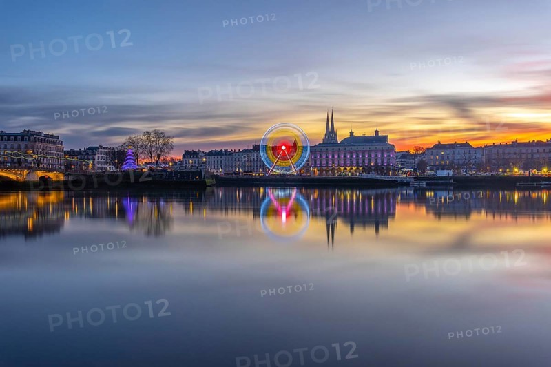 Bayonne, france, on christmas night experiencing a beautiful sunset reflecting upon the adour river, with festive lights illuminating the city buildings and a ferris wheel in motion