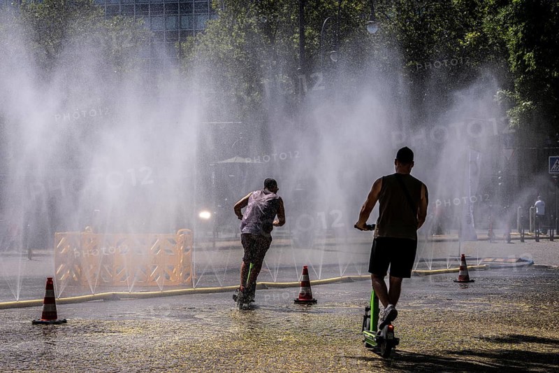 Cooling Cologne, campaign by the city of Cologne to cool down places exposed to heat, here Rudolfplatz in the city center, at just under 37°C during the day, a spray hose splashes fine water droplets that cool down the immediate area a few degrees, an action carried out when there is extreme heat stress, to adapt the urban climate to climate change, passers-by enjoy the cooling moment, plus tips on how to deal with extreme Hitze, North Rhine-Westphalia, Germany