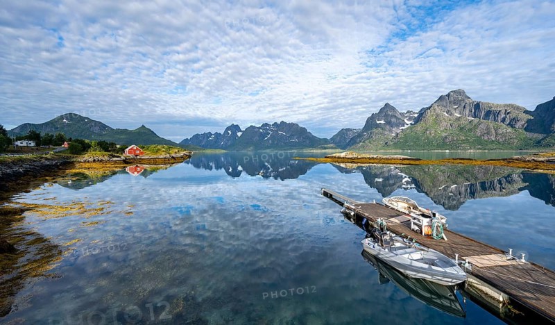 Red fishing hut on the shore and boats on a jetty, Rorbuer hut, picturesque fjord landscape at Raftsund fjord, mountains reflected in the water, Vesterålen, Norway