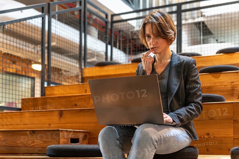 Focused young woman in jeans and blazer thinking deeply while working on laptop in modern coworking space