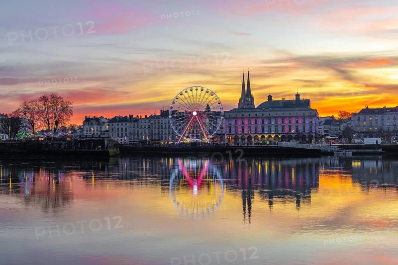 Bayonne skyline glowing at sunset with festive lights, ferris wheel and cathedral reflected in the calm adour river, winter dusk cityscape radiating holiday charm
