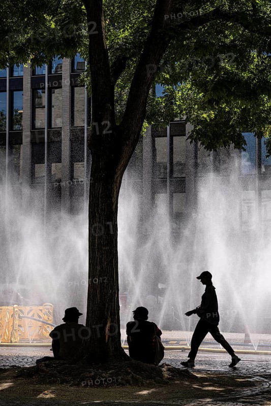 Cooling Cologne, campaign by the city of Cologne to cool down places exposed to heat, here Rudolfplatz in the city center, at just under 37°C during the day, a spray hose splashes fine water droplets that cool down the immediate area a few degrees, an action carried out when there is extreme heat stress, to adapt the urban climate to climate change, passers-by enjoy the cooling moment, plus tips on how to deal with extreme Hitze, North Rhine-Westphalia, Germany
