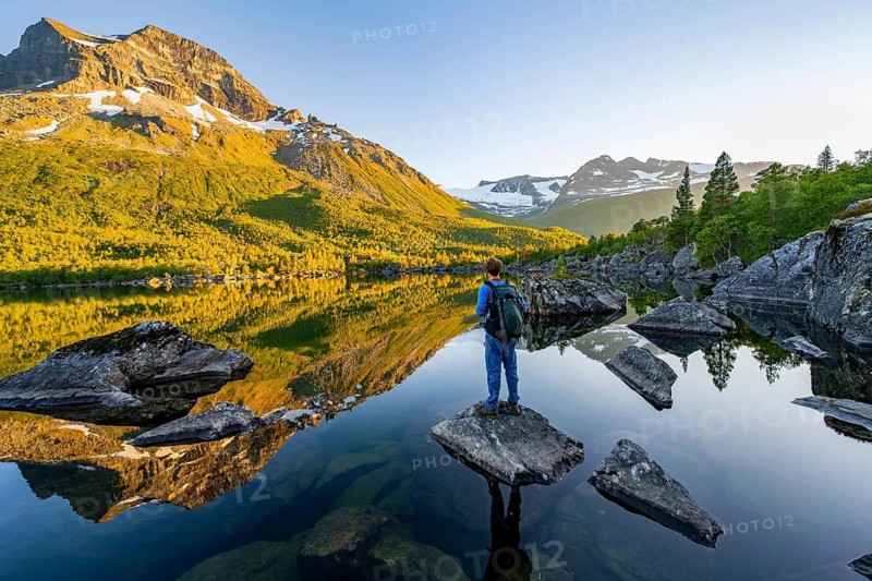 Young man standing on a rock in water, enjoying the landscape, picturesque mountain scenery in the Innerdalen high valley, mountain peaks reflected in Lake Innerdalsvatna, in the evening light at sunset, Trollheimen Mountain Area, Sunndal, Møre og Romsdal, Norway