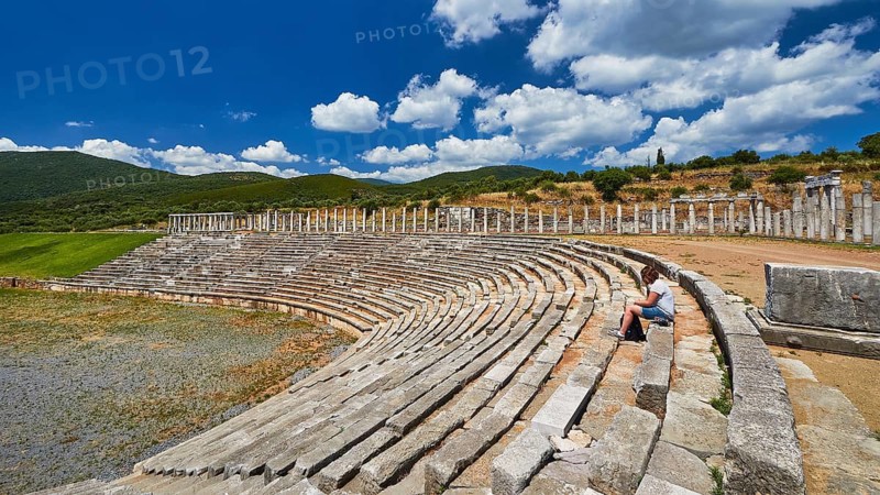 Person sitting in ancient ruins of a stadium under blue sky with clouds, Messene Stadium, Ancient Site, Archaeological Site, Messene, Messini, Messinia, Southwest of the Peloponnese, Odyssey, Odysseus, Ulysses, Movie, Filming Location, Matt Damon, Peloponnese, Peninsula, Greece